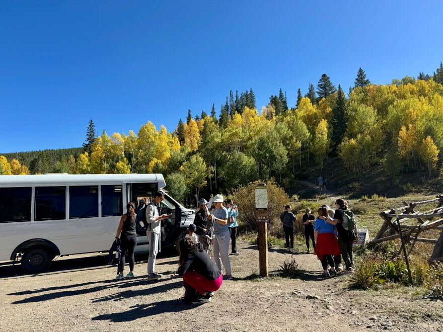 A shuttle bus drops off hikers in a dirt parking lot in front of a trail with yellow leaves