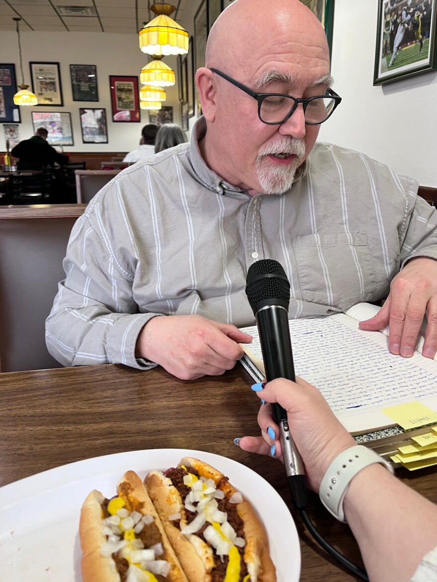 Flint native Tim Retzloff sits at a table at Sparty's Coney, reading from a stack of journals. April holds a microphone for him. A plate of coneys, one Detroit style and one Flint style, sits on the table in front of Tim.