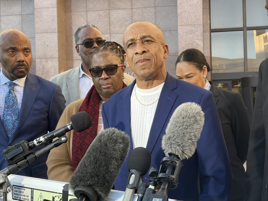 A man stands in front of microphones while speaking. A group of people stands behind him.