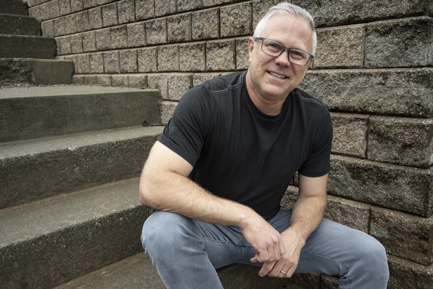A man wearing a black T-shirt and jeans sits on concrete steps near a brick retaining wall. He is gently smiling and looking at the camera.