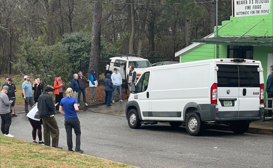 A line of people stand on the street in front of a white van and a green building.
