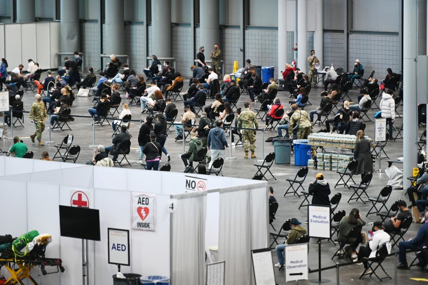 People move through a busy convention center (the Javits Center) to get vaccinated. Ther are walled off areas, as well as spaced out chairs where people sit and wait.