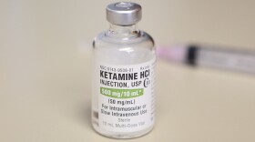A clear bottle with a label that reads "ketamine" stands on a white table.