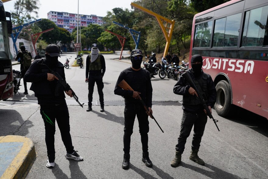 Three armed, masked men dressed in black stand guard on a street near a red bus, with additional masked individuals and motorcycles in the background. Trees and a colorful building are visible further back.