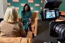 Marva Johnson sits in a chair speaking with Gina Jordan as a camera captures video of the interview.