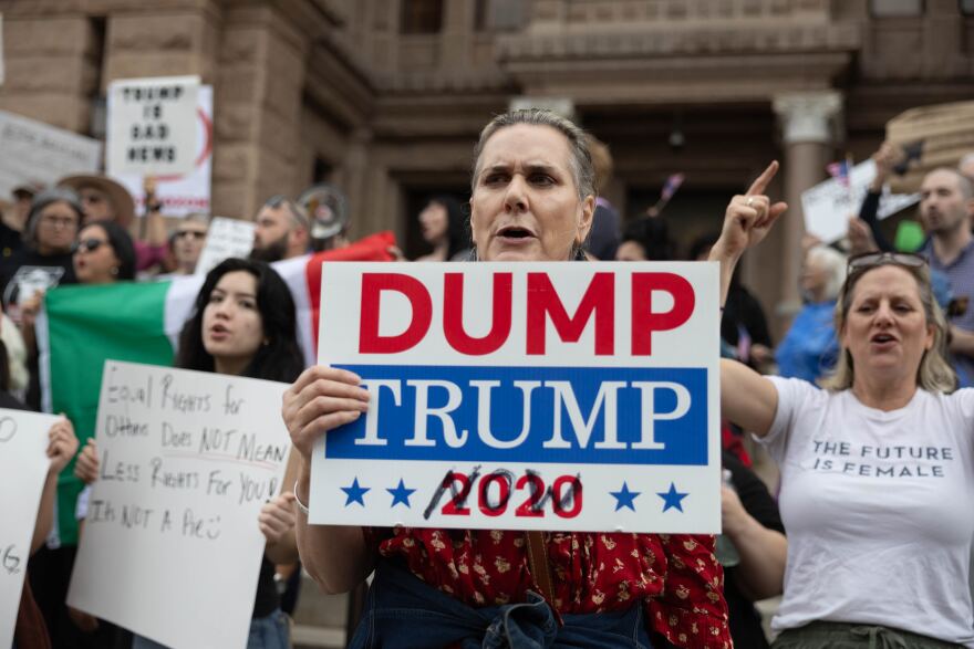 A protester holds a sign that says "Dump Trump Now," with the word "now" scribbled over the word "2020."