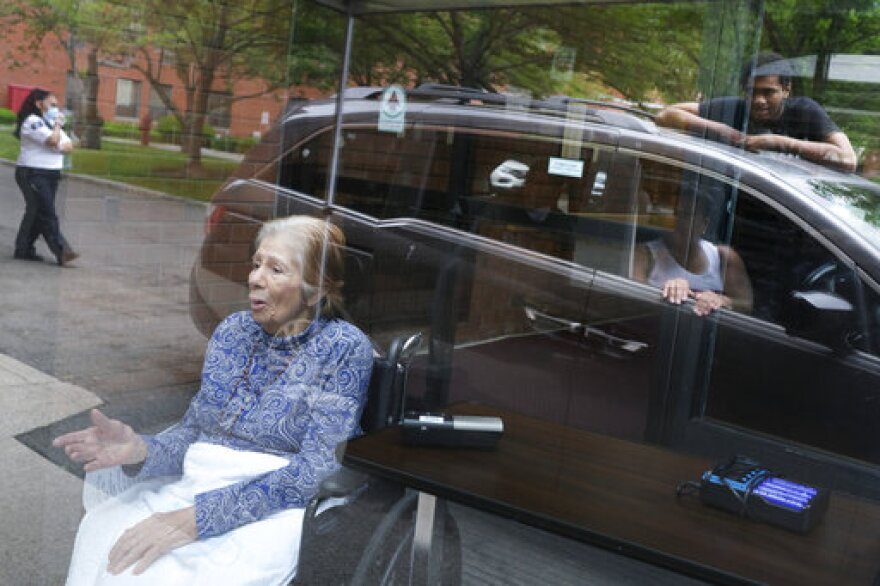 Gloria DeSoto, 92, bottom left, visits with her family, reflected in the glass in their car, from a window of the Hebrew Home at Riverdale, where she lives, in New York, Thursday, June 11, 2020. Her family was able to visit by a speaker while they stayed in their car and DeSoto stayed behind the glass of an entryway. They had not been able to visit since March 10, when many nursing homes began to shut down visitations for fear of spreading the coronavirus. The Hebrew Home at Riverdale, home to 700 residents, just started these non-contact visitations and will be scheduling them seven days a week to help families and residents reconnect. (AP Photo/Seth Wenig)