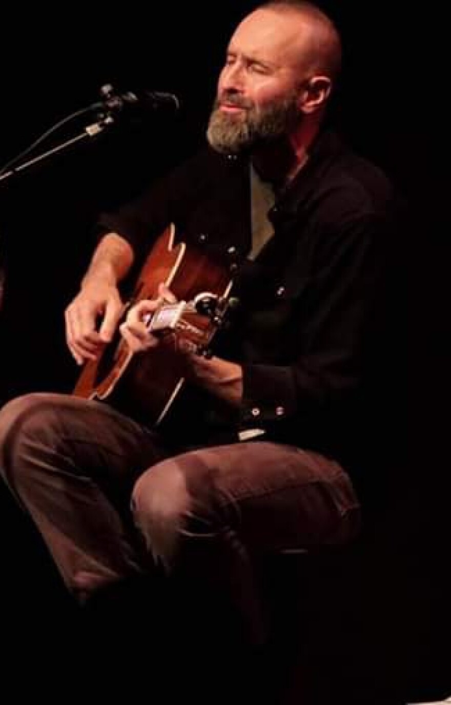 Cecil sits on a stool playing his guitar and sings into a mic.