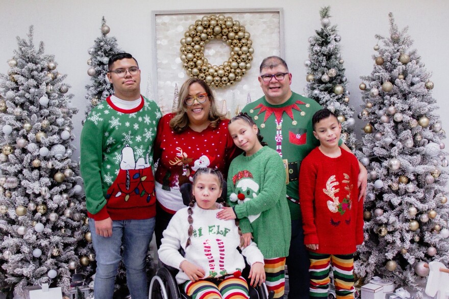A Christmas-themed family portrait, with a mom, dad and four kids posing in red and green Christmas sweaters with patterns like elf's stockings and reindeer antlers. One of the kids, in front, uses a wheelchair.