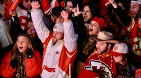 A crowd of people wearing mostly heavy, red winter clothing are bunched together. One person wearing a white jacket with a "Chiefs" logo is raising his hands in celebratory fashion. Others are cheering around him.