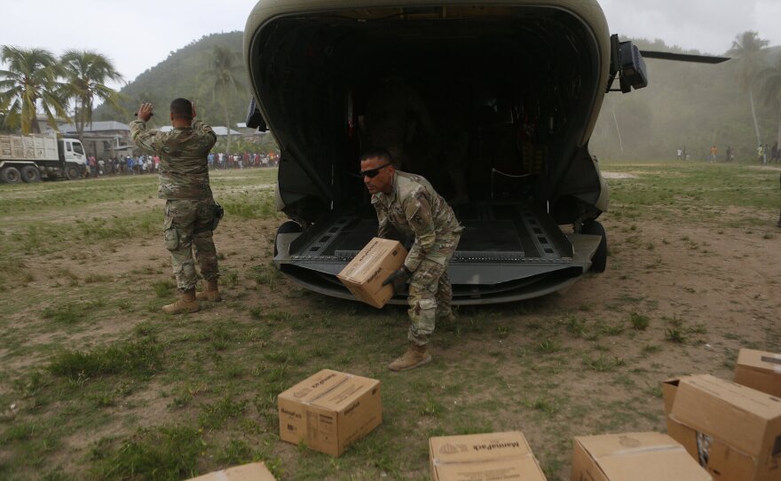 Men in U.S. military fatigues unload cardboard boxes from a helicopter in a field of grass and dirt.