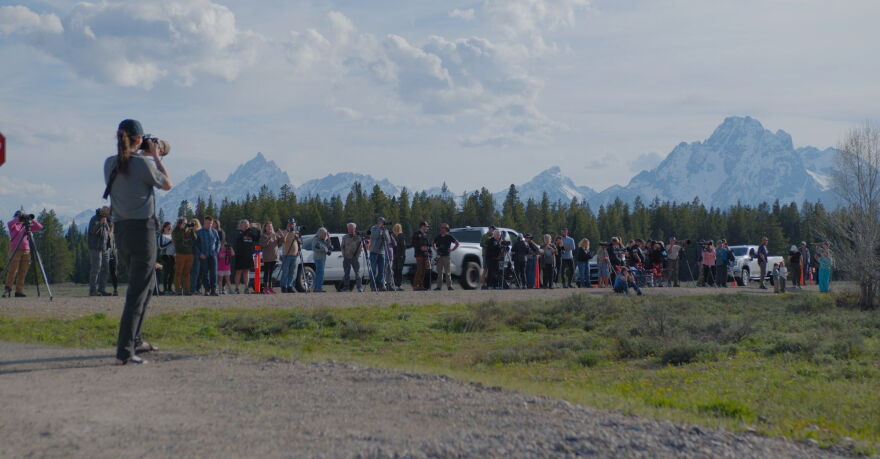 A group of people, many with cameras, gather along a road beneath hovering mountains.