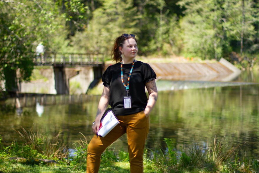 Woman, holding a notebook, stands at the edge of a lake.
