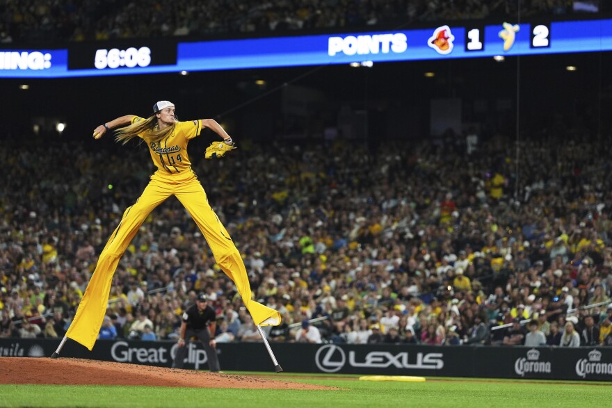 a man with long hair years a yellow uniform and throws a baseball pitch while on stilts