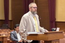 A bearded man in a blazer and tie speaks at a podium in a courtroom, with another man sitting behind him.
