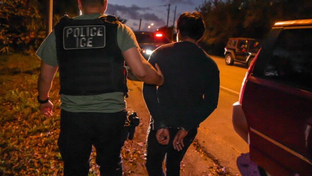 An immigration officer wearing a black flack jacket that says "POLICE ICE" on the back holds he arm of someone who appears to be a man and whose hands are cuffed behind his back. The two walk amidst dark-colored cars on a road at night.