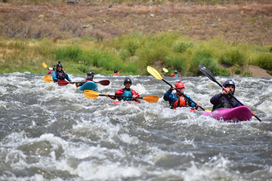 A group of kayakers in helmets and life jackets paddle through choppy whitewater on the Klamath River near the former Iron Gate Dam site.