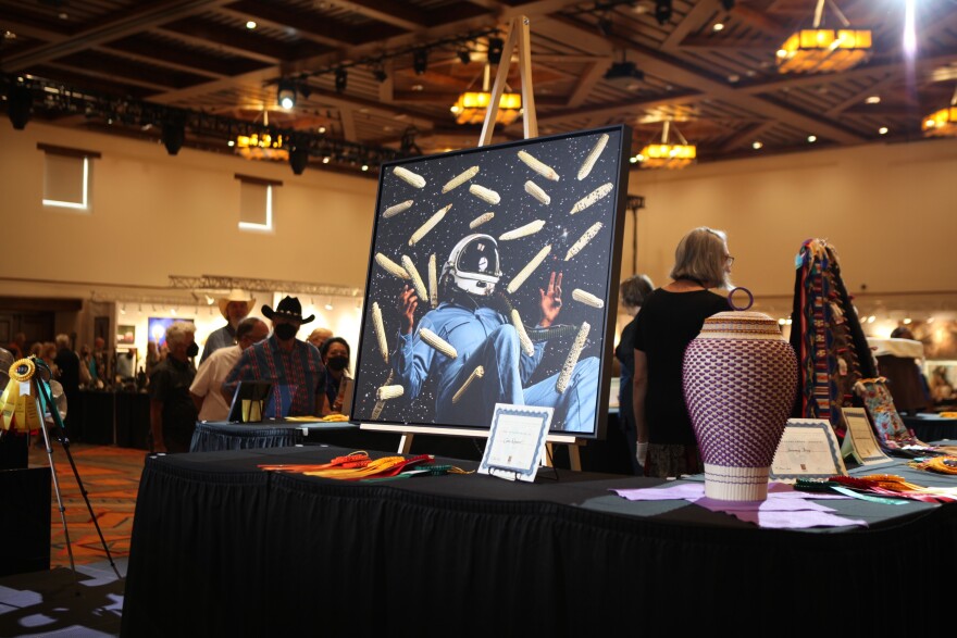 A large print of a photograph of an astronaut in a starry sky surrounded by corn, with a purple basket alongside, in a large hall with artworks and viewers