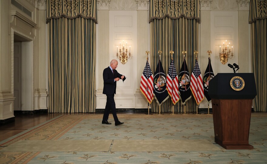 A man with gray hair checks his watch as he walks across a carpeted floor towards a wooden podium, with a row of American and presidential flags against the wall.