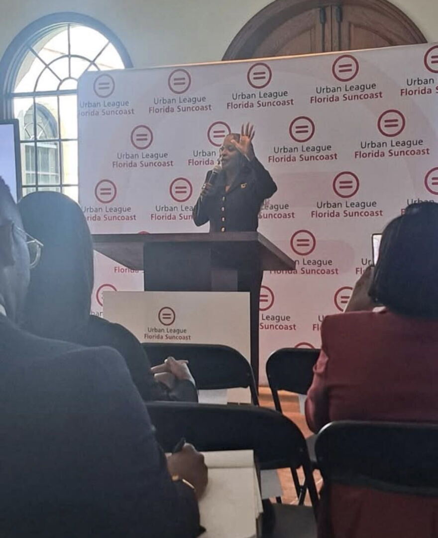 A women on a stand is speaking to people sitting in chairs with a Urban League of Florida Suncoast banner behind her