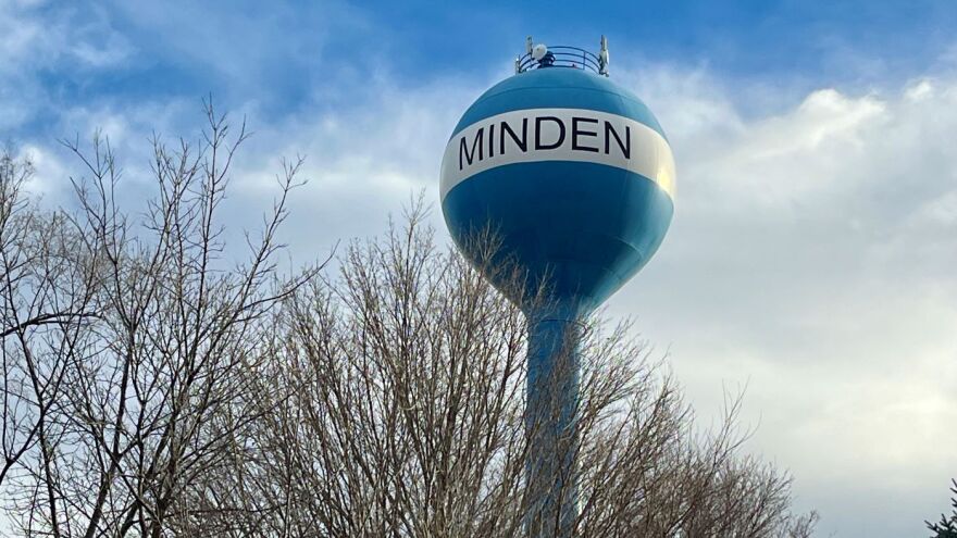 Picture of a blue water tower between trees without leaves. The water tower says "Minden" in black letters