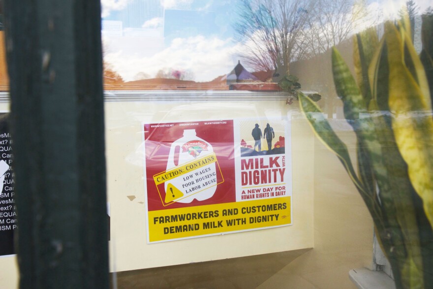 A photo looking throw window glass at a red and yellow poster that reads Milk with Dignity, a new day for human rights in dairy, farmworkers and customers demand milk with dignity, caution consumer, low wages, poor housing, labor abuse.