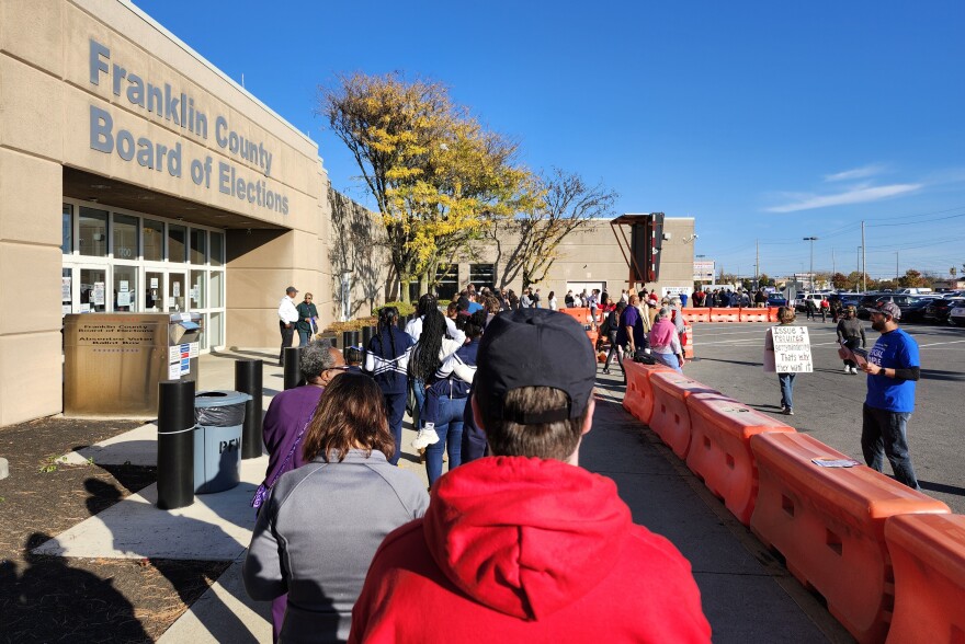 A long line of people seen from behind wait alongside a large building. The building is labeled "Franklin County Board of Elections." To the right, a barrier separates the line from a few stray people wearing signs or passing out pamphlets.