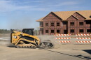 A yellow bulldozer uses a brush attachment to sand down poured concrete in front of a framed condo structure 