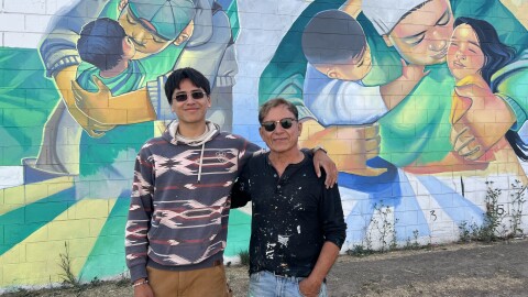 two men stand in front of a colorful mural 