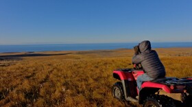 A man wearing a winter coat sits on a four wheeler and looks out through binoculars across a wide plain that gently descends to the outside of Savoonga, Alaska.