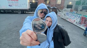 Two women in light blue hoodies stand in a parking lot holding a sterling silver, heart-shaped case that says "bang bang" on the lid.