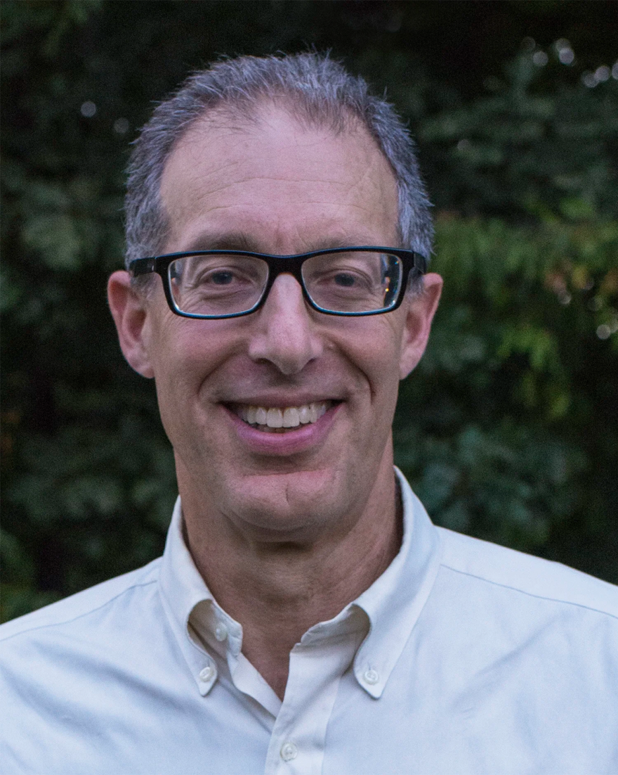 A man with gray hair and black glasses smiles at the camera, a blurry green background behind him.
