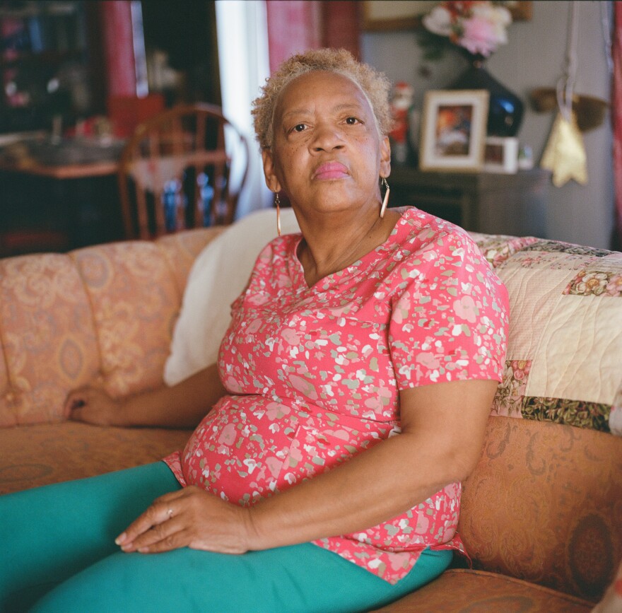 Older Black woman with a pink blouse sitting on a couch and looking straight into the distance
