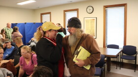 Ken Bradshaw, a man in his fifties with a long gray beard and spectacles, holds a crying blonde girl in her 20s. They're standing in a busy town hall meeting room. 