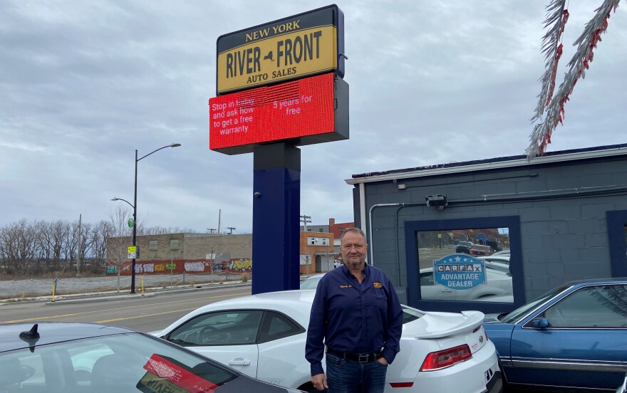 A man in jeans and dark blue button-up shirt stands in front of a white car and blue car. A large sign behind him reads "River Front Auto Sales."