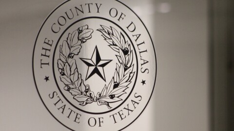 A transparent version of the Dallas County seal on a glass door. The seal includes the words "The County of Dallas - State of Texas" as well as a star in the center of two curved branches of leaves.