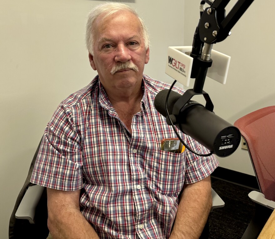 Elderly man with white hair and a mustache sitting in a chair, wearing a plaid shirt. He's in front of a microphone on a boom arm. The setting appears to be a radio station studio.