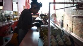 A lone figure of a woman is silhouetted against a large window in a restaurant space. She appears to be working on pastries set out on the counter.