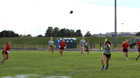 High school girls playing flag football on a grass field at a school. Several players are wearing orange and black jerseys. The others are wearing gray and green jerseys. 