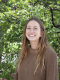 Emily Calhoun stands outside in front of a green, leafy tree. She is smiling widely.