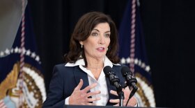 Gov. Kathy Hochul, wearing a blue suit and white blouse, speaks at two microphones.