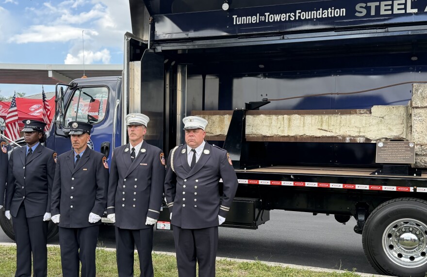 Four firefighters in navy blue uniforms stand at attention near a truck with a side panel open that shows a horizontal beam from the World Trade Center