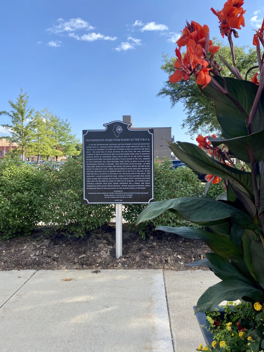 A planter of summer flower in full bloom foregrounds a square, brown marker installed on the edge of a municipal parking lot.