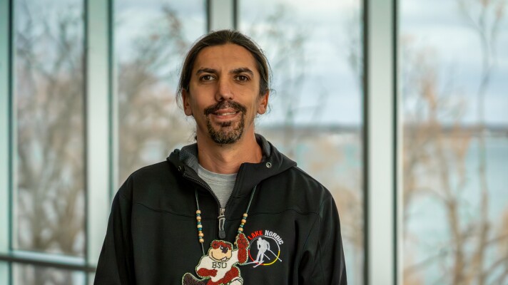 A man in a black jacket and BSU beaded necklace stands in front of windows overlooking a lake in winter.