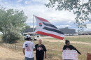 A boy in an American flag t-shirt holds up an Arapaho flag. He stands next to an older woman in a black t-shirt and another woman in a colorful skirt holding up a protest sign.