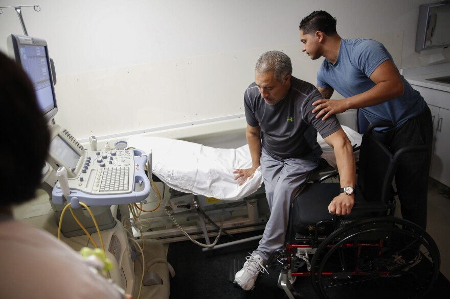 Joel Ramirez is helped onto a bed to receive an ultrasound with the aid of Francisco Guardado, his home health care giver, during a visit to the hospital in Rialto, Calif.