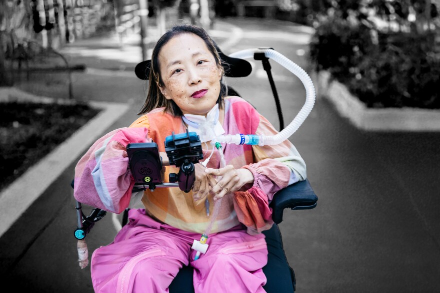 a woman in brightly colored, loose-fitting clothes, with a half-smile and brightly colored lipstick, sitting in a wheelchair with a breathing tube going from her neck to the back of the chair
