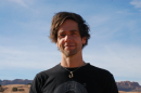 A man with medium-length brown hair stands outside. He wears a black shirt and a hook necklace. He looks at the camera and smiles. 