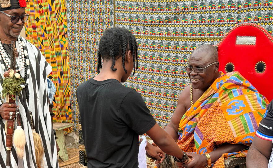 A young man kneels in front of an older man who is wearing a colorful robe.