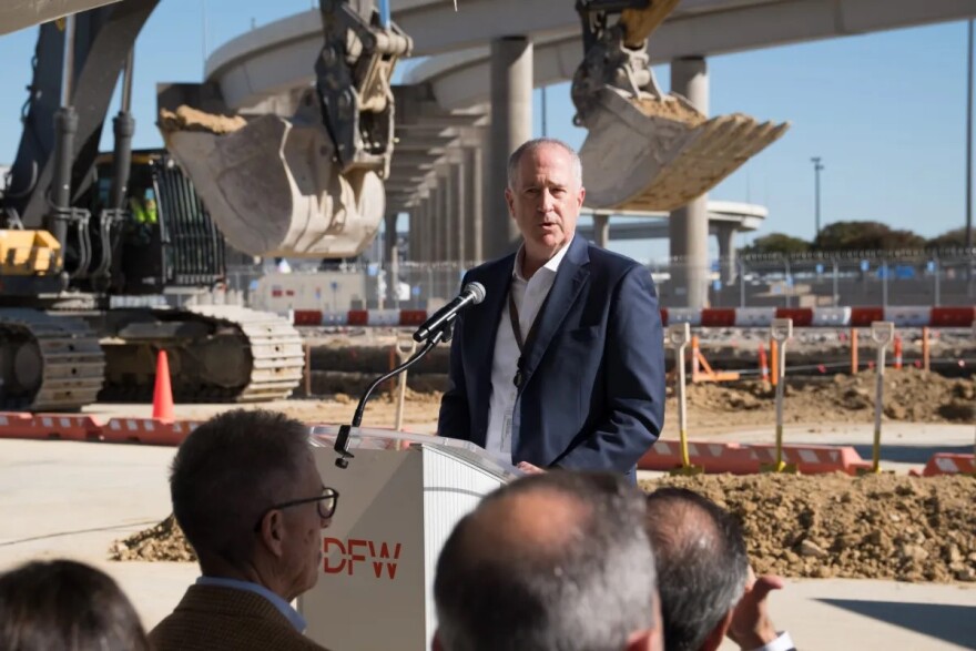 A man stands at a lectern with a microphone in front of a construction site. A group of people face him to listen.
